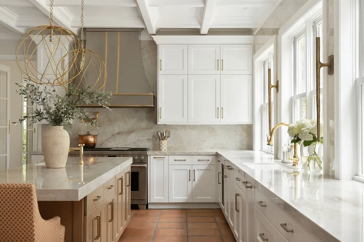 Bright white kitchen with marble countertops, gold hardware, and large windows; spherical pendant lights over an island.