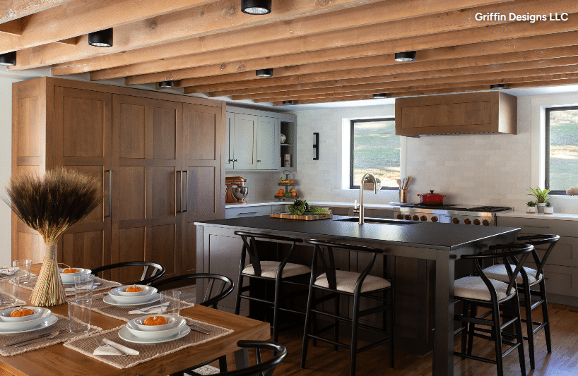 Open-concept kitchen with a large island, dark wood cabinets, stainless steel range, and a dining table set for six under exposed wooden ceiling beams.