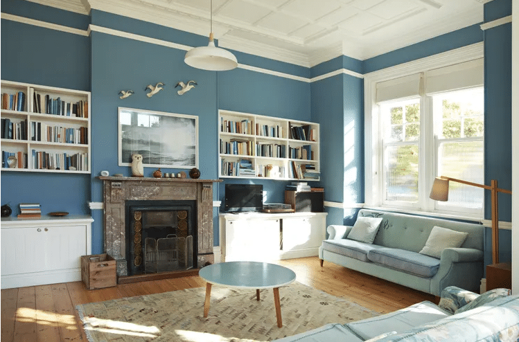 Bright blue living room with white built-in bookshelves, brick fireplace, and light wood floors; pastel sofa and round coffee table in center.