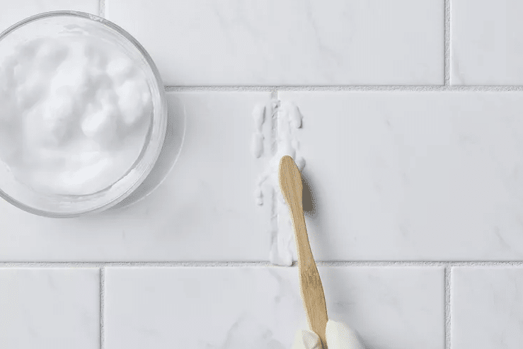 Wooden spatula smoothing white caulk along a grout line on white tiled wall, bowl of caulk nearby.