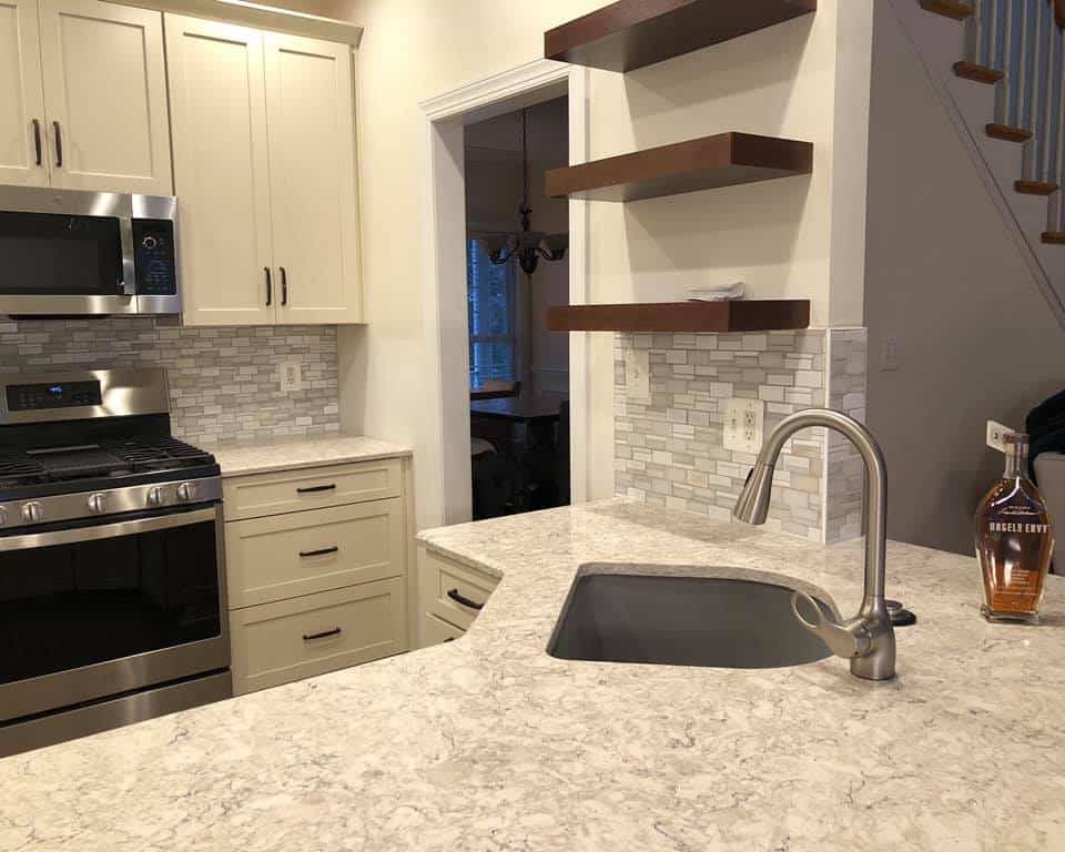 Modern kitchen with an island sink, stainless steel stove and microwave, cream cabinets, and floating wood shelves above a tiled backsplash.