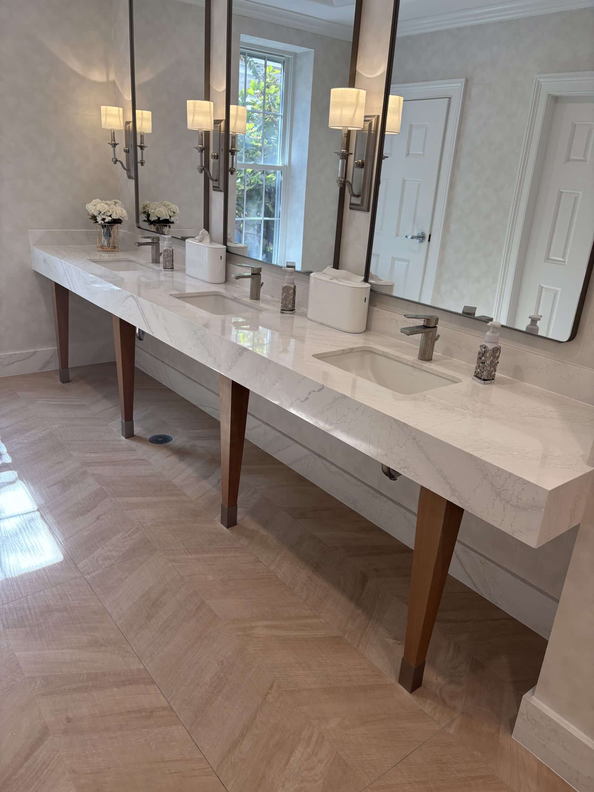 Double vanity with marble countertop, two sinks, tall mirrors, wall sconces, and decorative flowers in a vase.