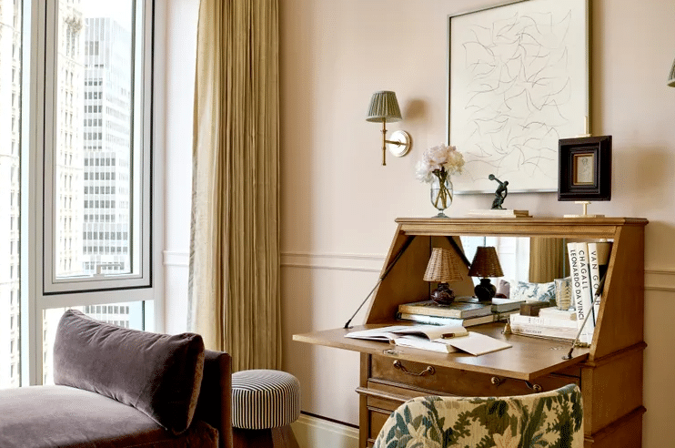 Wood roll-top desk open with books, lamp, and vase on top in a sunlit beige living room by a large window showing city skyscrapers.