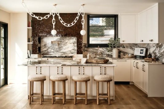 Bright modern kitchen with marble backsplash and island, four wooden stools, white cabinets, brick accent wall, and a large window.